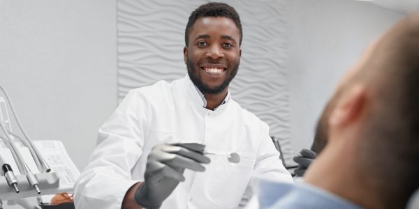 Selective focus of cheerful male dentist in process of curing teeth in dentist office. Professional doctor looking at camera and laughing while patient lying on dentist chair. Concept of stomatology.